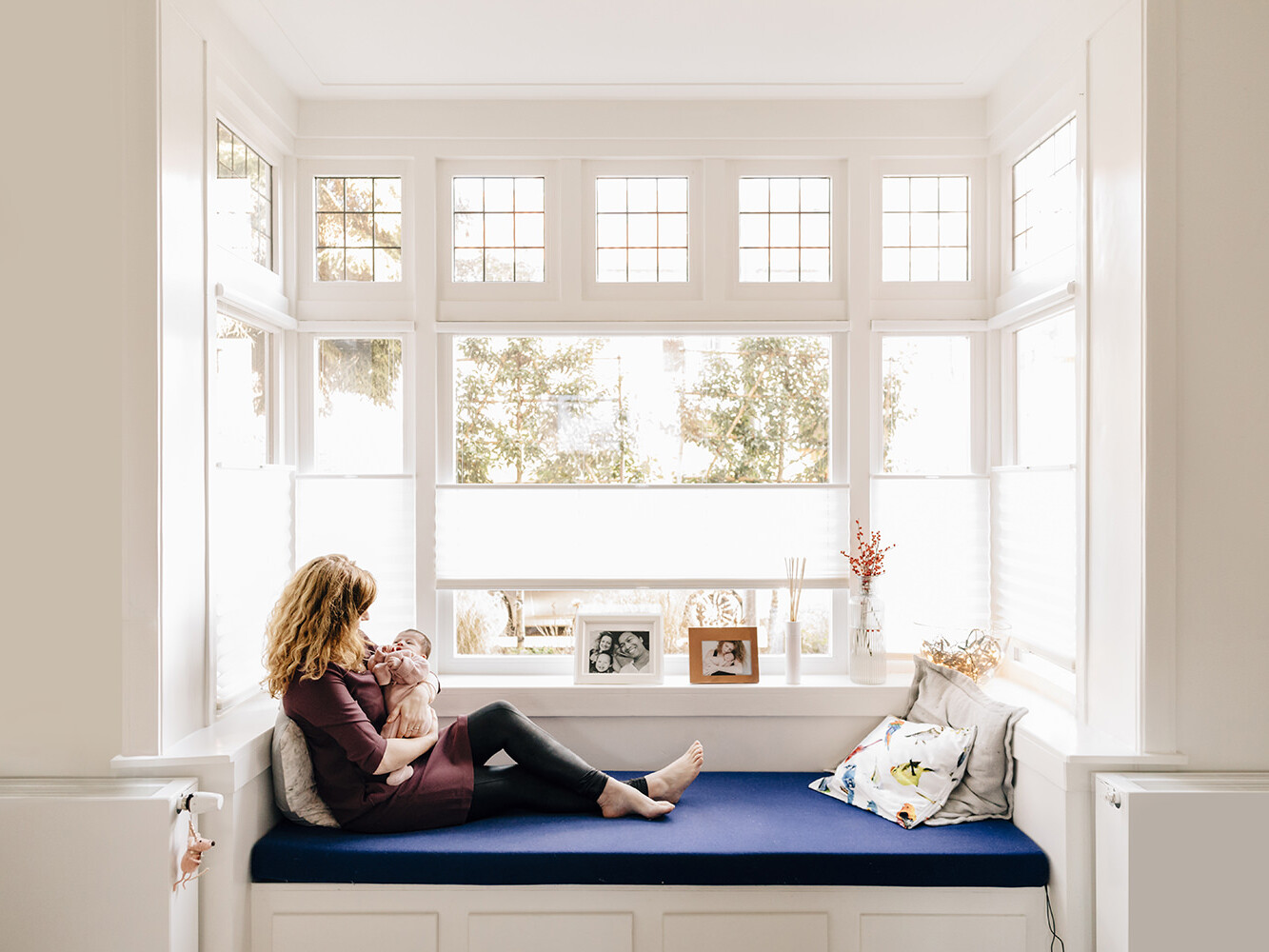 foto van een moeder die met haar baby knuffelt op een bank voor een hele mooie jaren 30 raam tijdens een gezinsfotoshoot aan huis