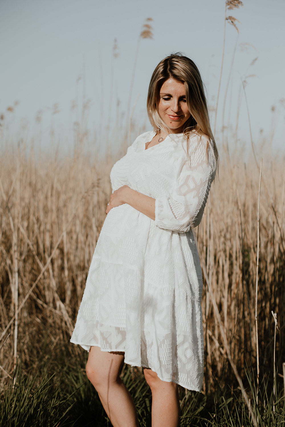 foto van een mooie jonge zwangere vrouw met lang blond haar en een witte jurk tijdens een zwangerschapsshoot buiten op locatie