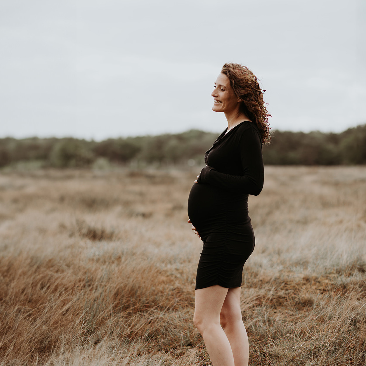 foto van een mooie jonge zwangere vrouw met krullend haar en een zwarte jurk tijdens een zwangerschapsshoot op de hei