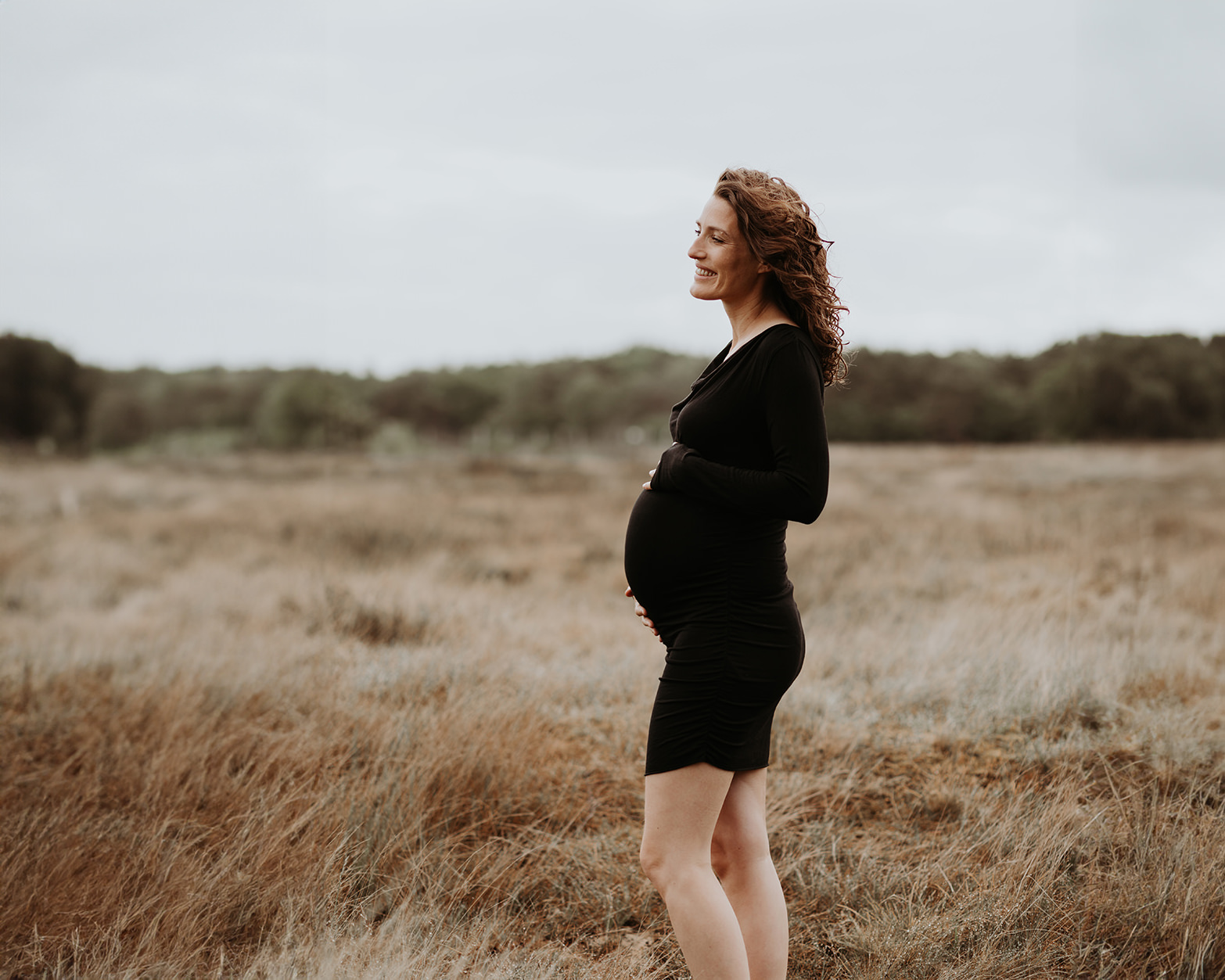 foto van een mooie jonge zwangere vrouw met krullend haar en een zwarte jurk tijdens een zwangerschapsshoot op de hei