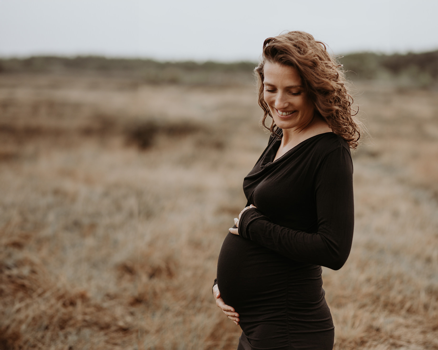 foto van een mooie jonge zwangere vrouw met krullend haar en een zwarte jurk tijdens een zwangerschapsshoot op de hei