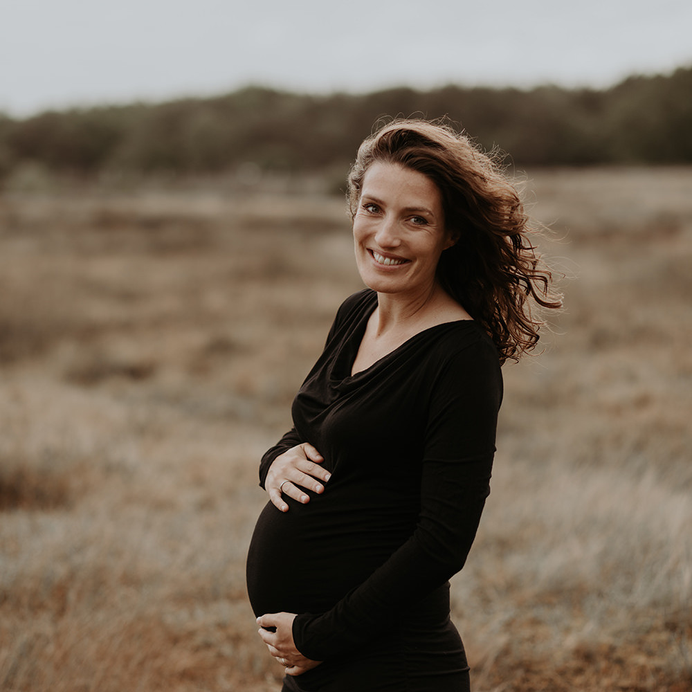 foto van een mooie jonge zwangere vrouw met krullend haar en een zwarte jurk tijdens een zwangerschapsshoot op de hei