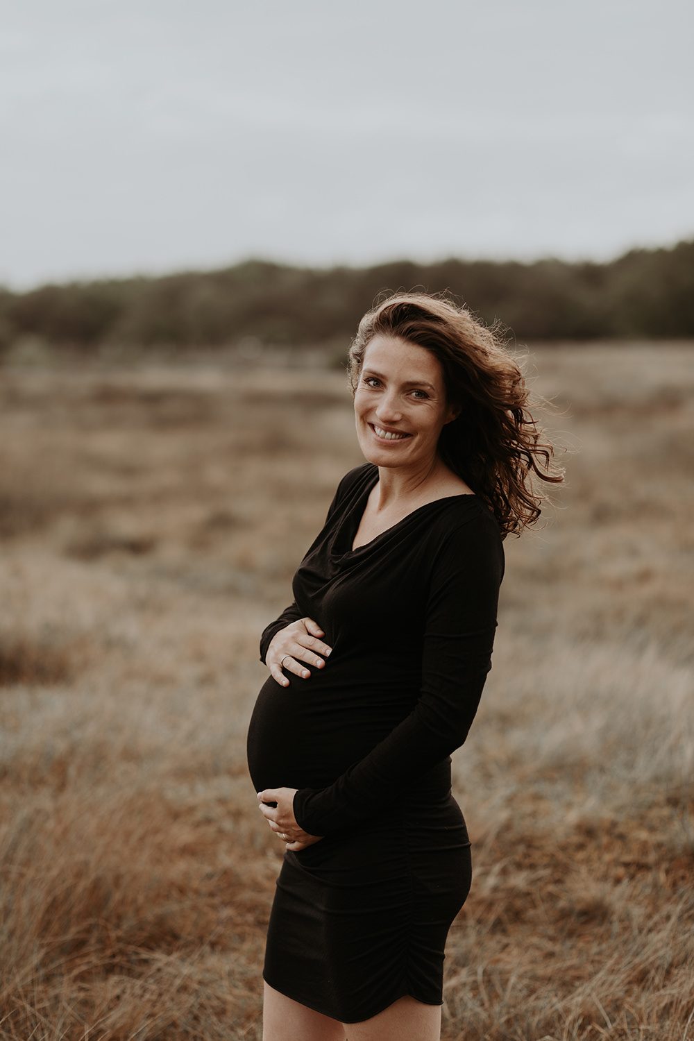 foto van een mooie jonge zwangere vrouw met krullend haar en een zwarte jurk tijdens een zwangerschapsshoot op de hei