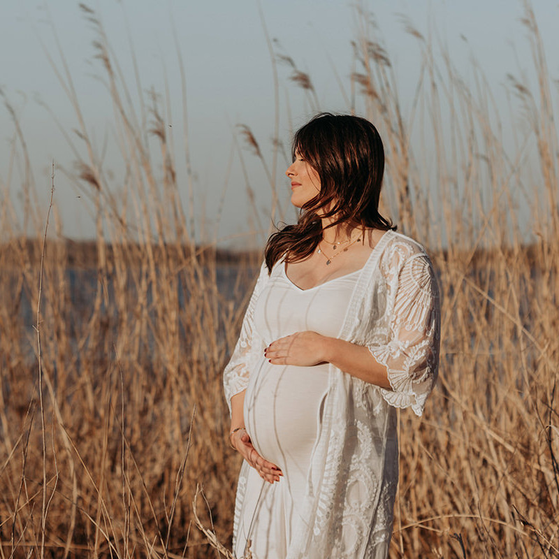 foto van een mooie jonge zwangere vrouw met lang bruin haar en een witte jurk tijdens een zwangerschapsshoot op het strand