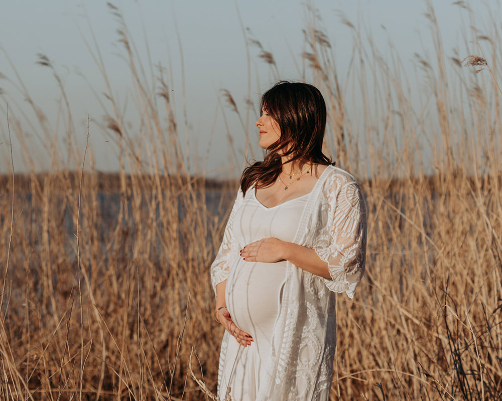 foto van een mooie jonge zwangere vrouw met lang bruin haar en een witte jurk tijdens een zwangerschapsshoot op het strand